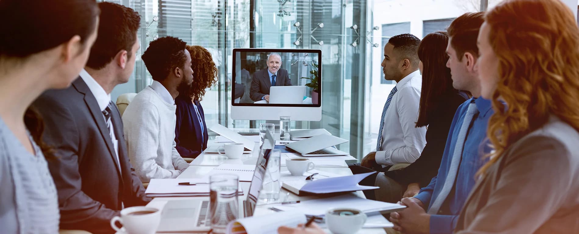 A group of professionals sit around a conference table watching a video call on a large screen, where a man appears to be speaking during a virtual meeting.