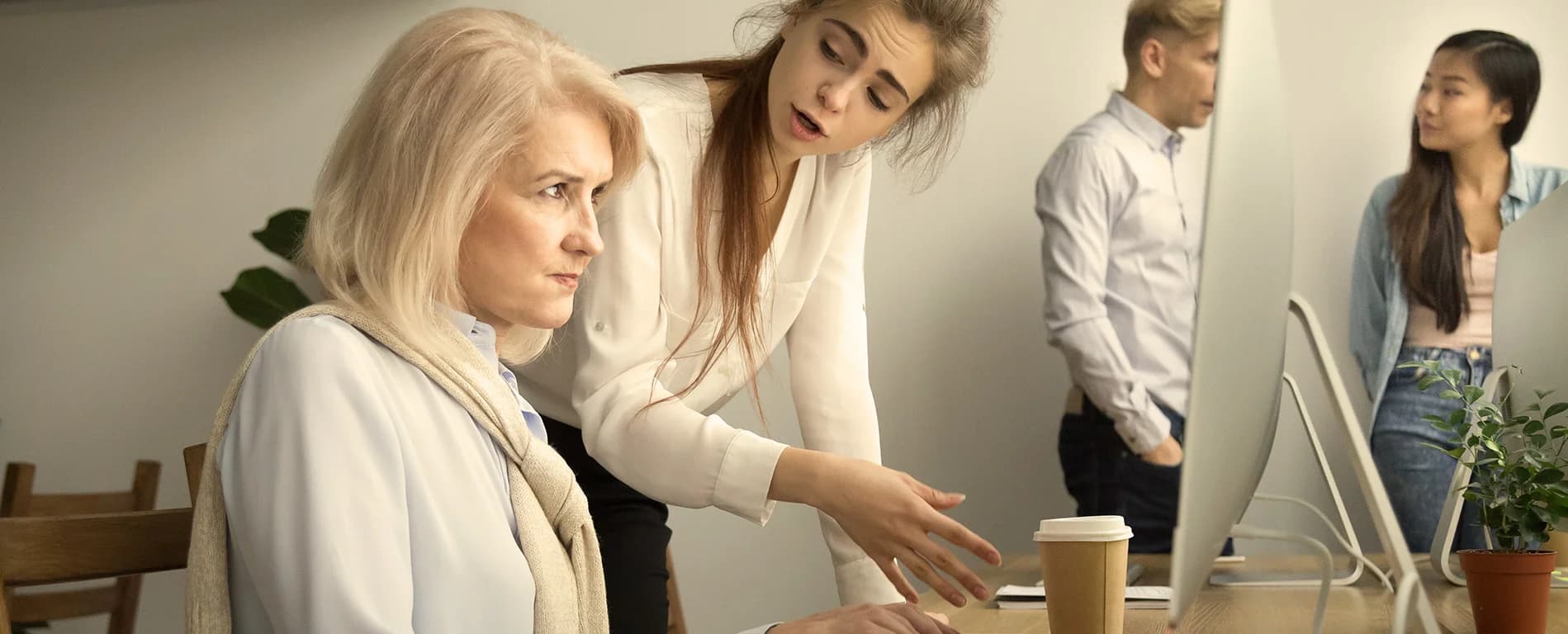 An older woman sits at a desk looking at a computer screen while a younger colleague leans over and gestures as if explaining something, with two other coworkers talking in the background.