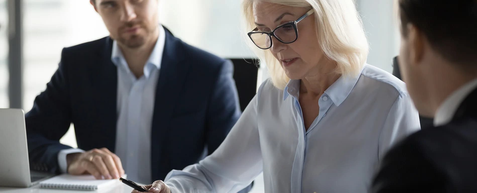 An older woman wearing glasses reviews and signs documents at a table while two colleagues sit nearby, discussing the paperwork.