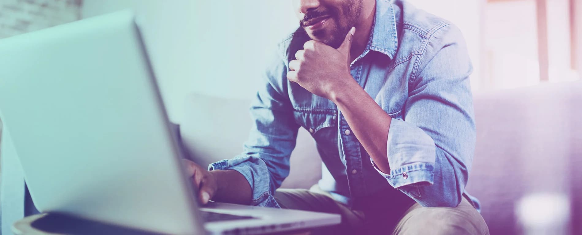 A man in a denim shirt sits on a couch using a laptop, resting his chin on his hand while looking thoughtfully at the screen.