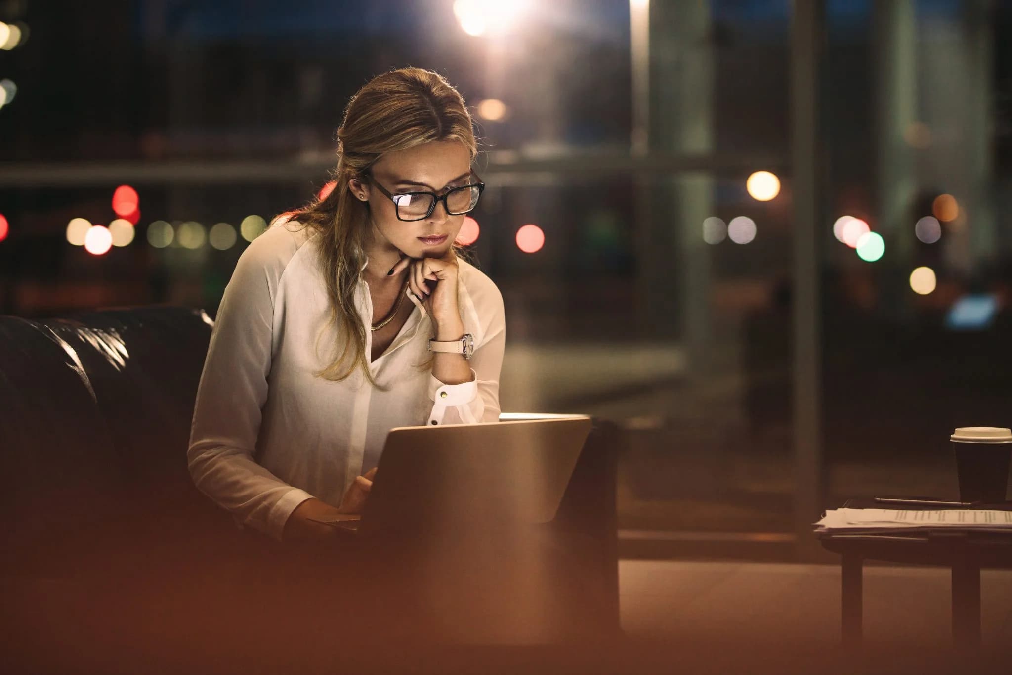 A woman wearing glasses sits indoors at night, focused on her laptop, with city lights visible through large windows in the background and a coffee cup on the table beside her.