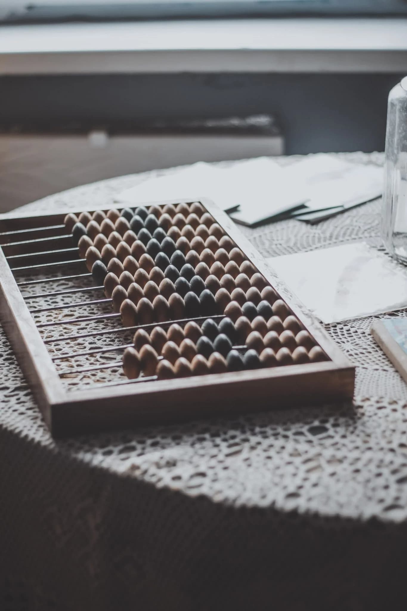 A wooden abacus with dark and light beads rests on a lace-covered table, with papers and a glass bottle nearby.