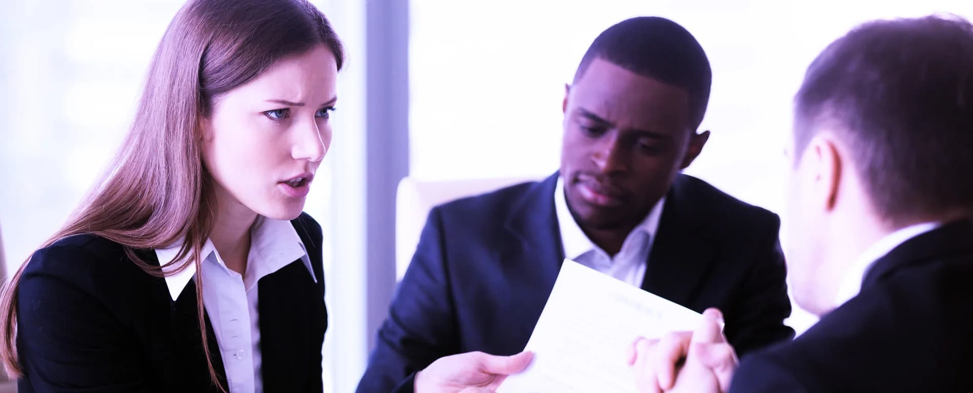 A businesswoman speaks firmly while holding a document, as two colleagues in suits listen during a discussion in an office setting.