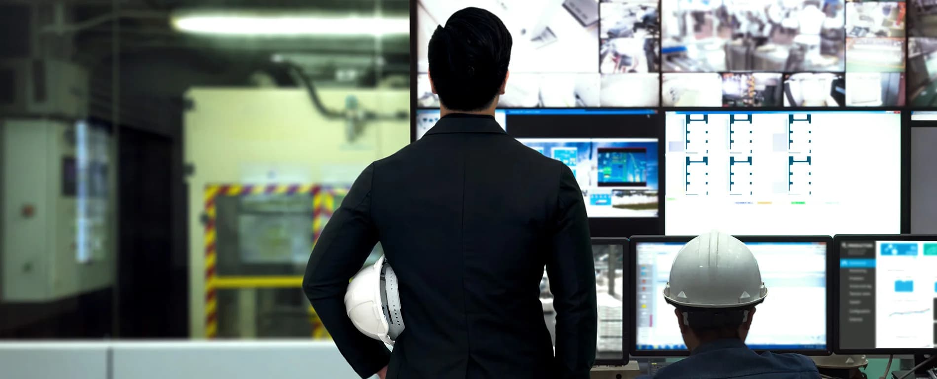 Two professionals monitor multiple large screens displaying surveillance footage and system data in an industrial control room, with one holding a safety helmet.