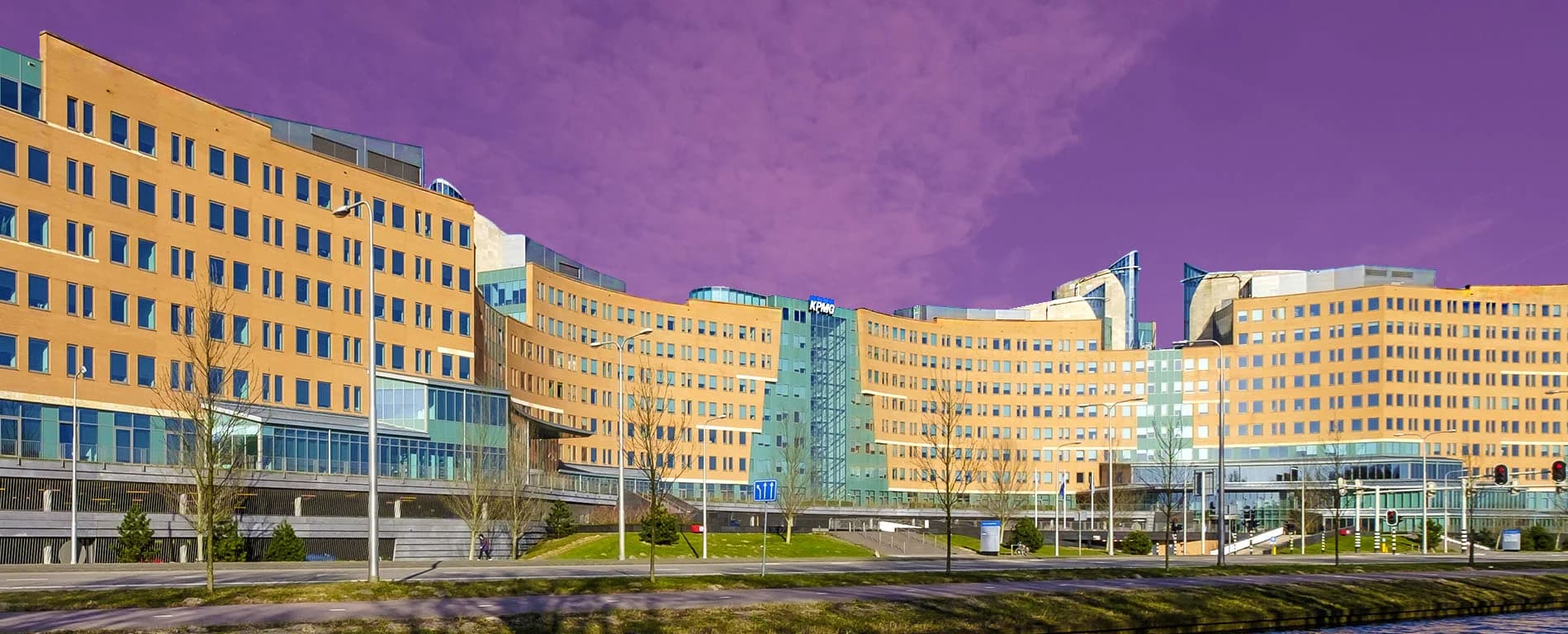 A large modern office complex with curved façades and many windows stands beside a roadway and green space under a purple-tinted sky.