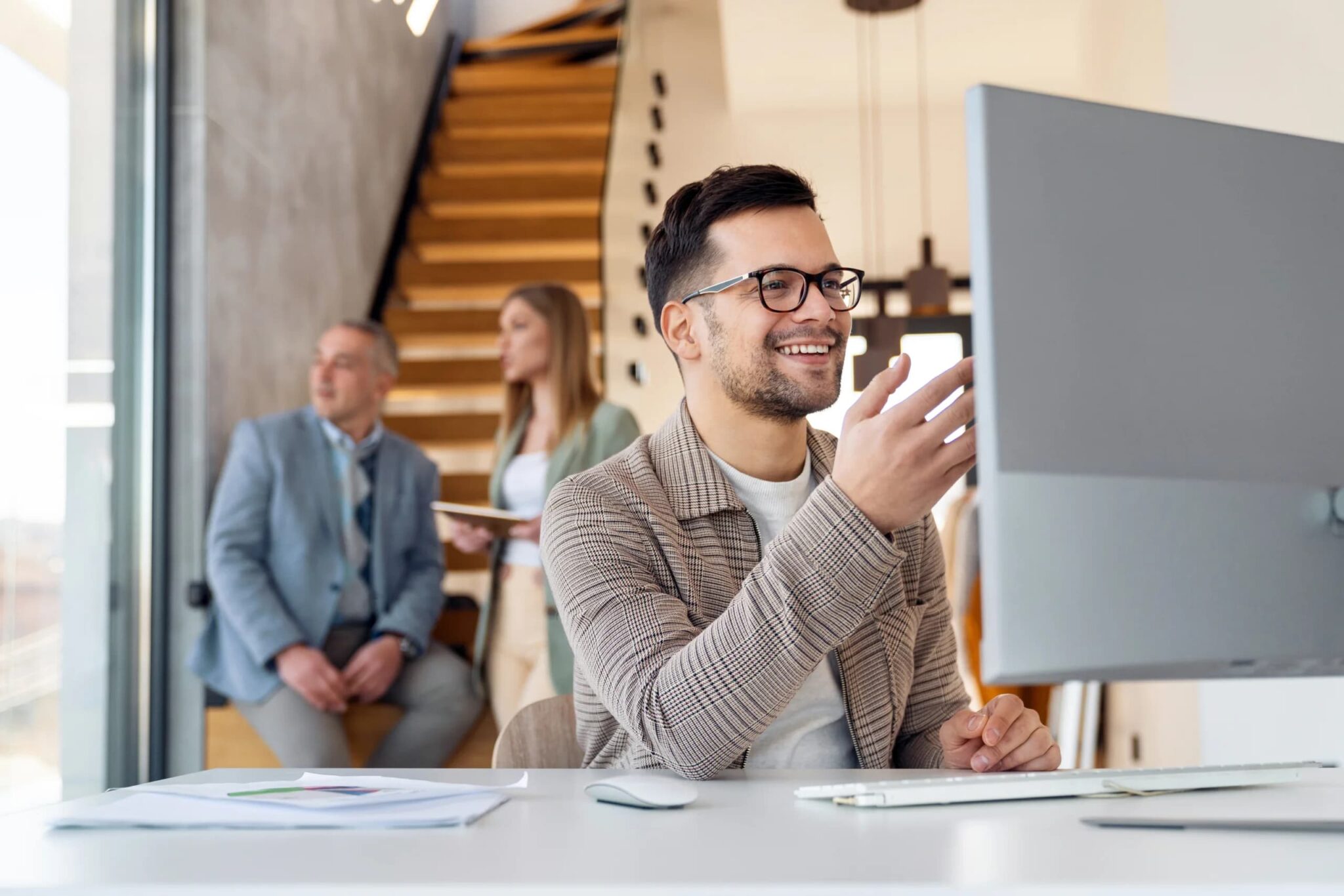 A smiling man wearing glasses works at a desktop computer in a modern office, while two colleagues stand and talk in the background near a staircase.