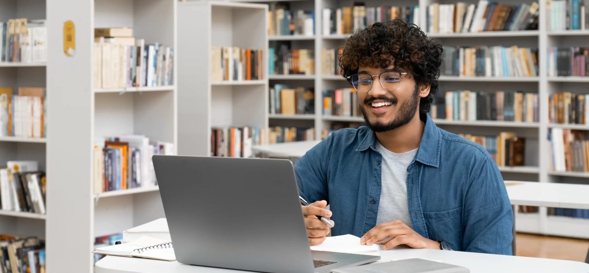 A smiling young man with curly hair and glasses sits at a table in a library, using a laptop and holding a pen with notebooks open in front of him, surrounded by bookshelves.