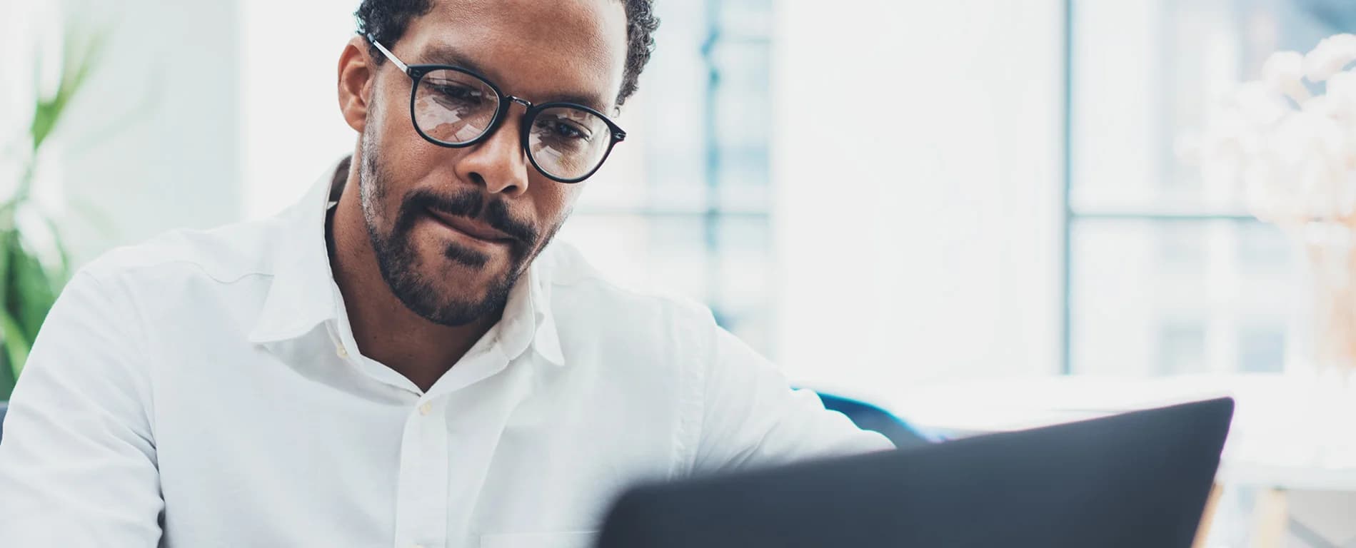 A man wearing glasses and a white shirt looks focused while working on a laptop in a bright office setting.