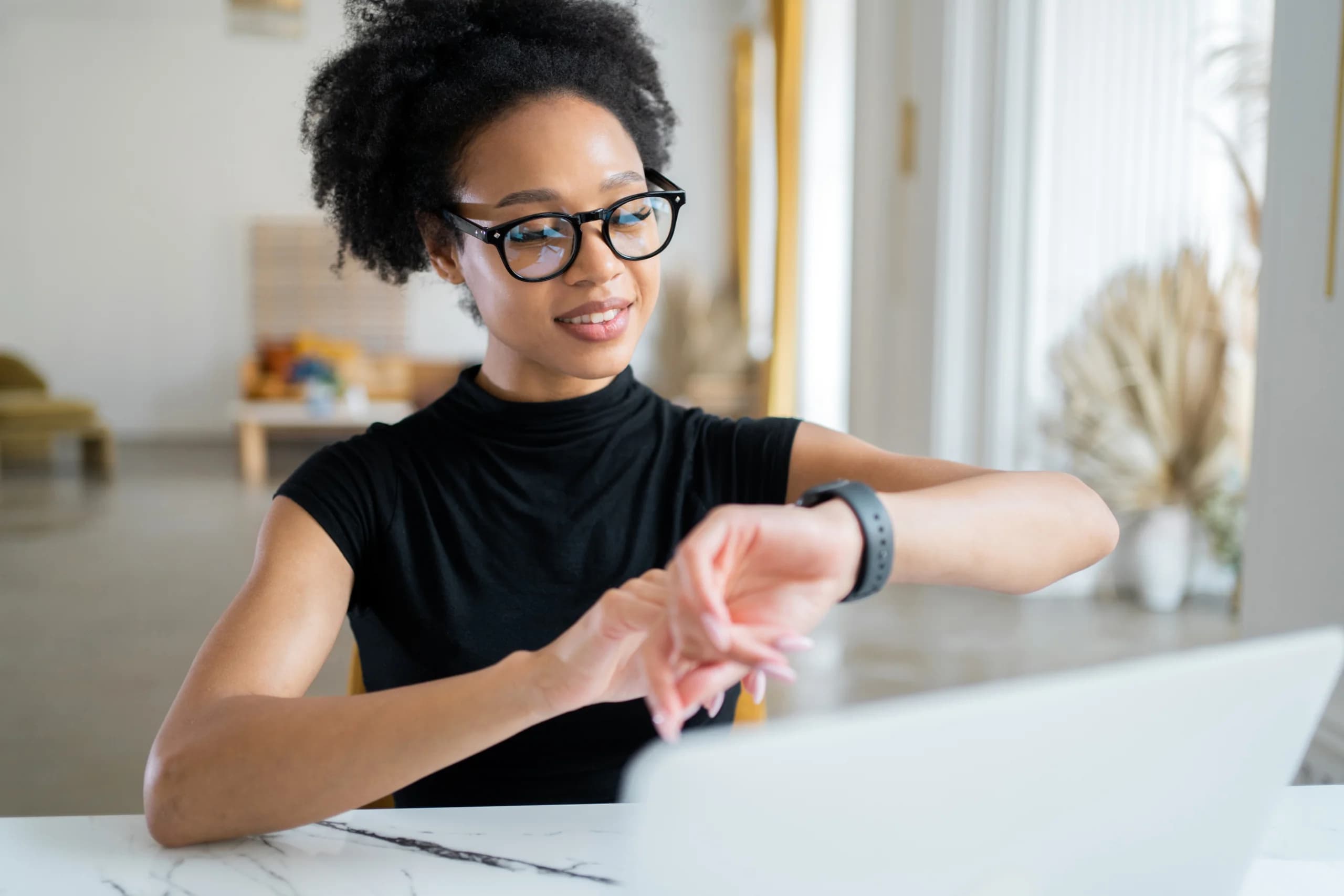 Woman wearing glasses checking her smartwatch while sitting at a desk with a laptop in a bright home office.