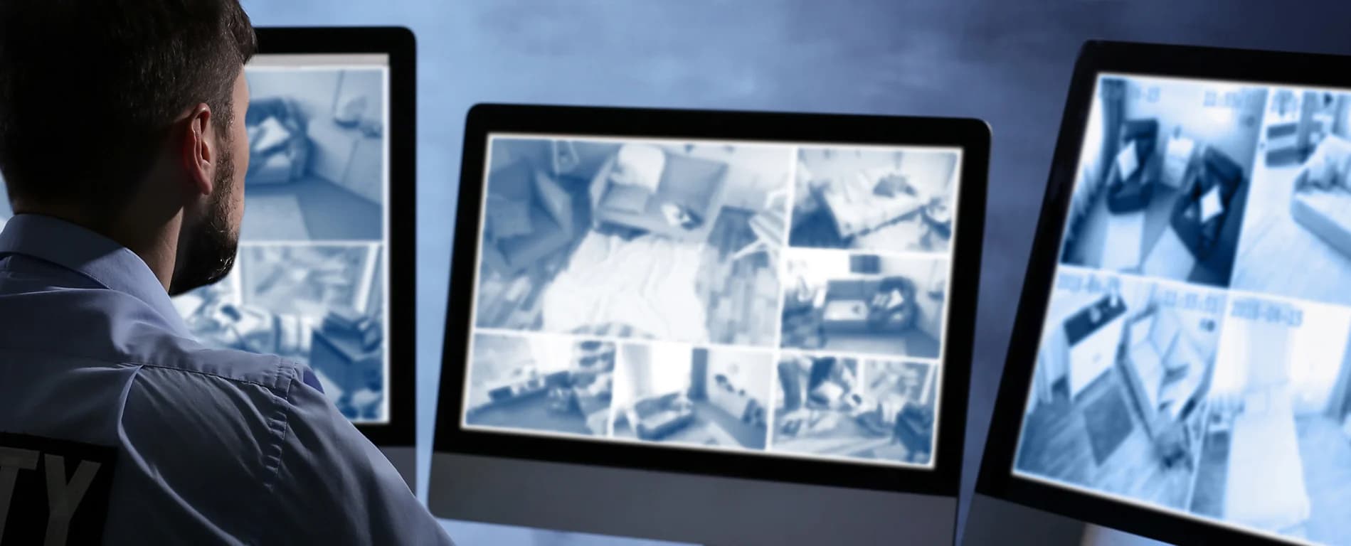 Man monitoring multiple security camera feeds on computer screens showing different indoor surveillance views.