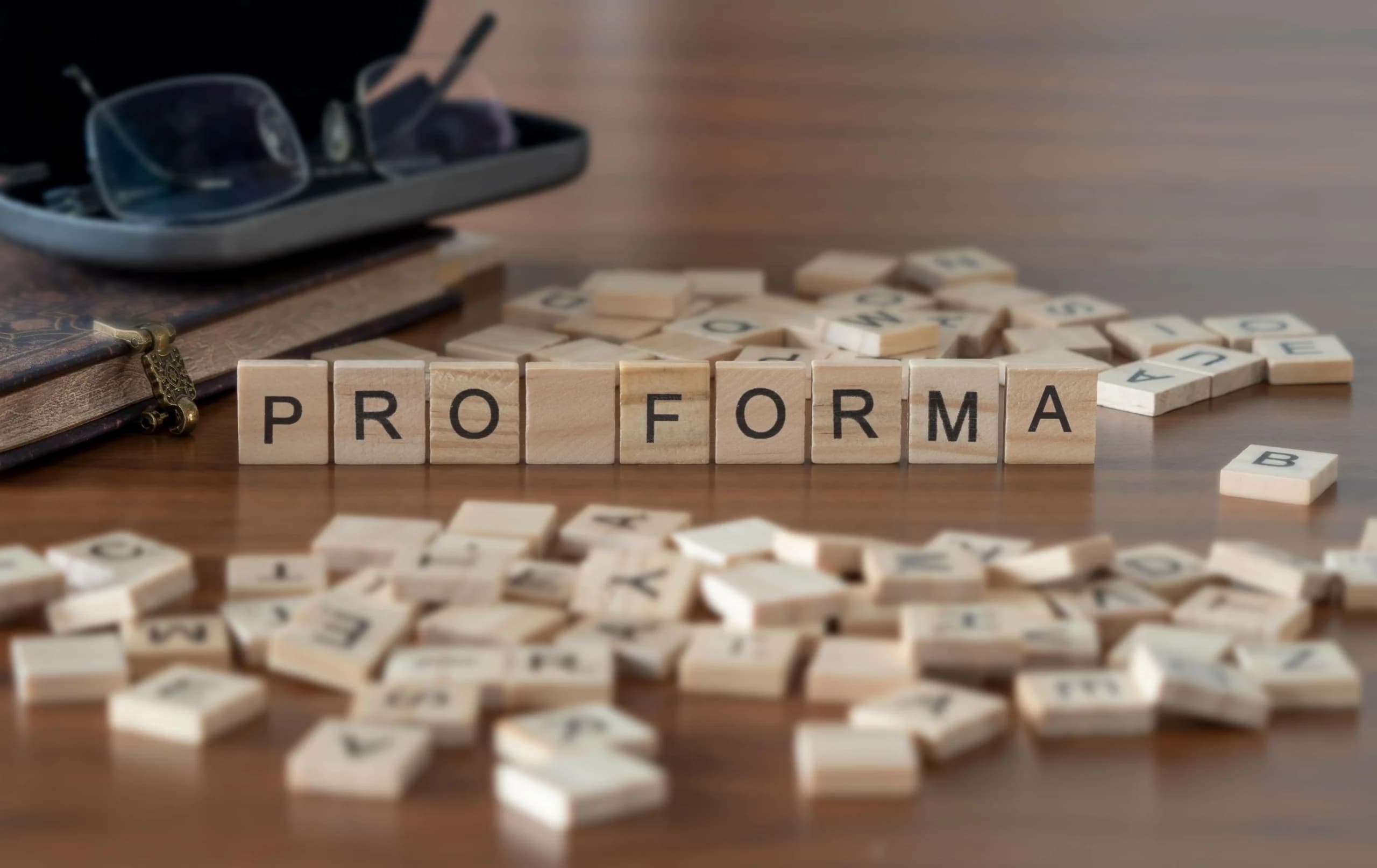 Wooden letter tiles spelling “PRO FORMA” on a desk, surrounded by scattered tiles, with a notebook and glasses in the background.