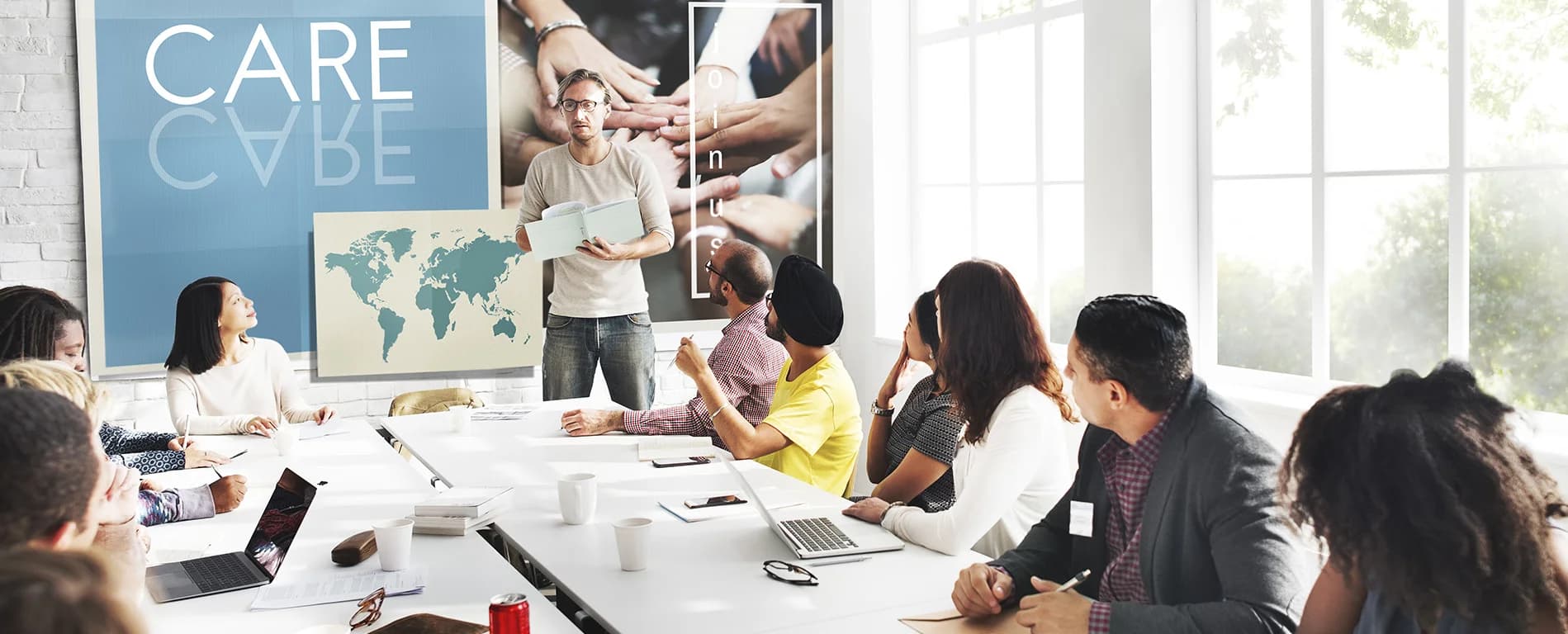 Man presenting to a diverse group in a meeting room with “CARE” displayed on the wall and teamwork visuals in the background.