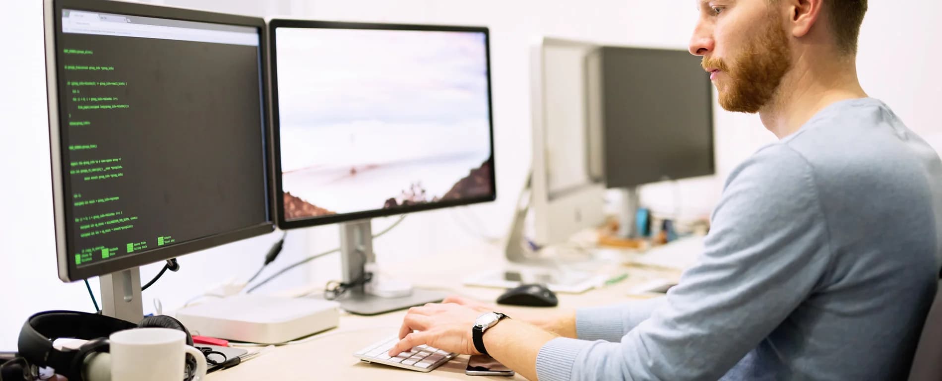 Man coding at a desk using dual monitors displaying programming code in an office environment.