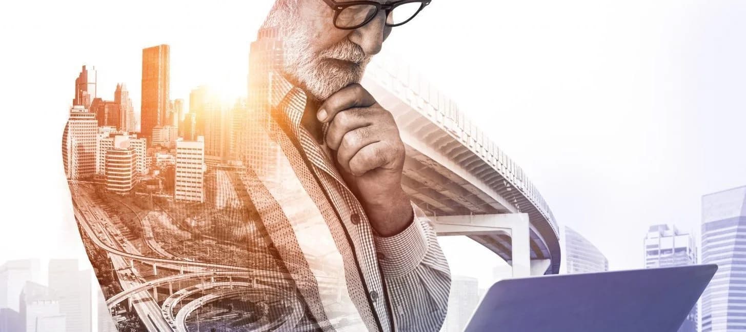 Double exposure of a thoughtful businessman using a laptop, blended with a cityscape and highway infrastructure, symbolizing urban business and strategic thinking.