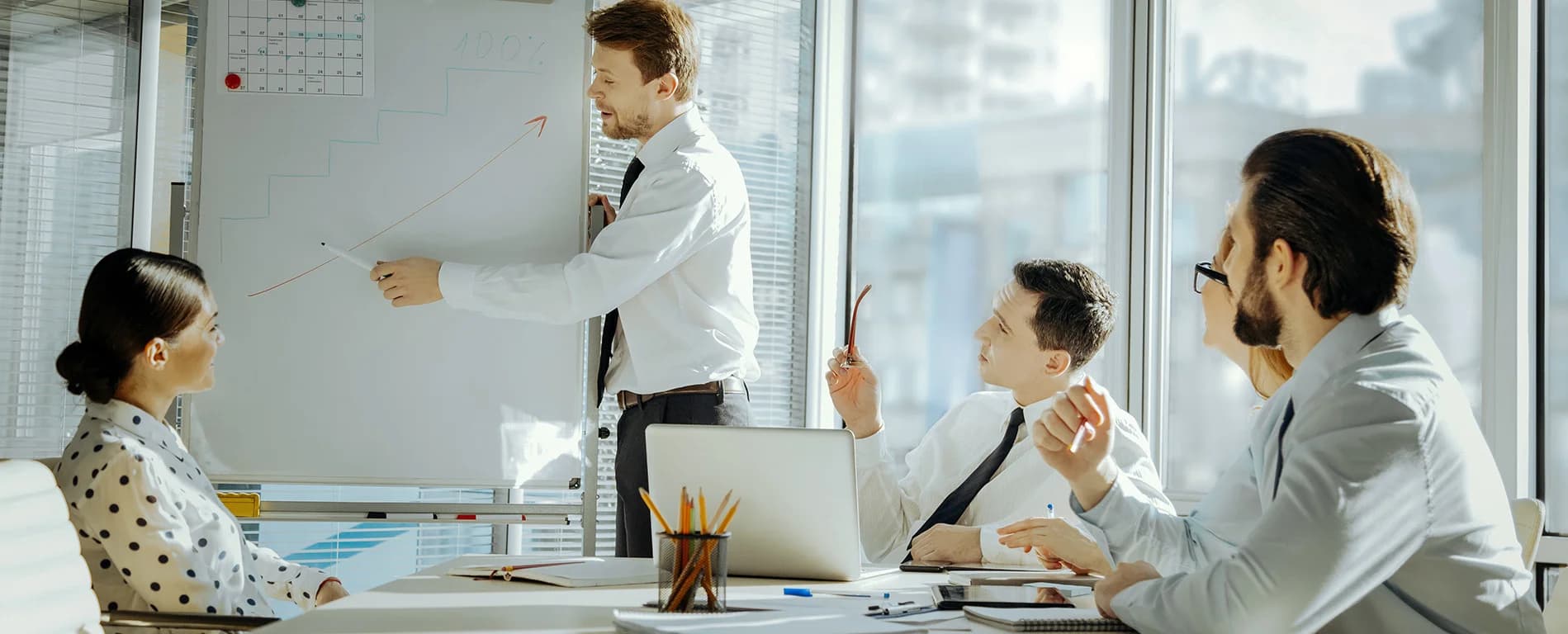Man presenting a rising graph on a whiteboard to colleagues during a business meeting in a bright office.