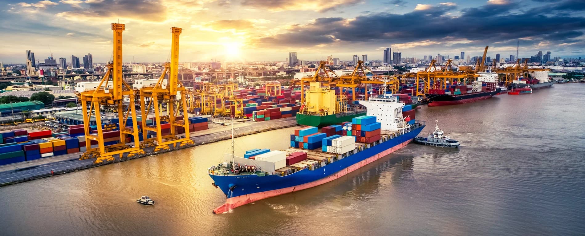 Container ship loaded with cargo at a busy port with cranes and stacked containers under a sunset sky.