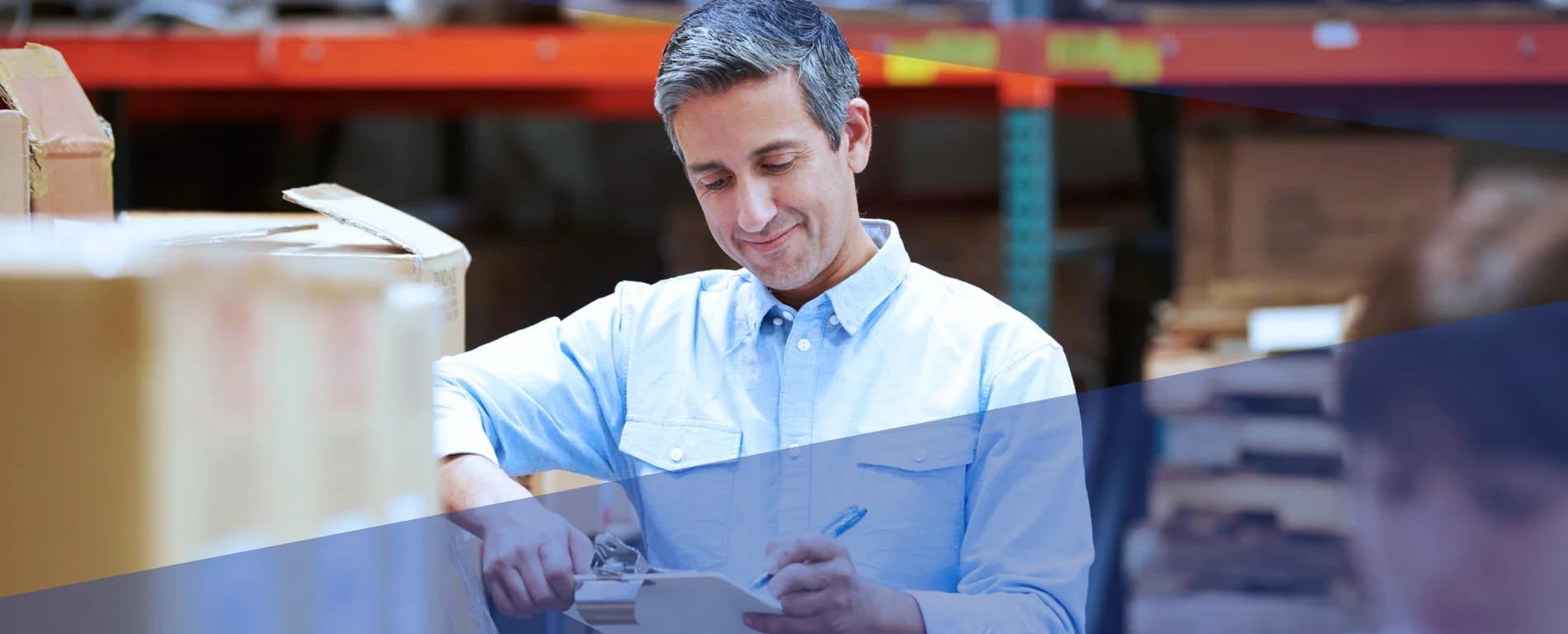 Warehouse worker checking inventory and writing on a clipboard next to stacked boxes in a storage facility.