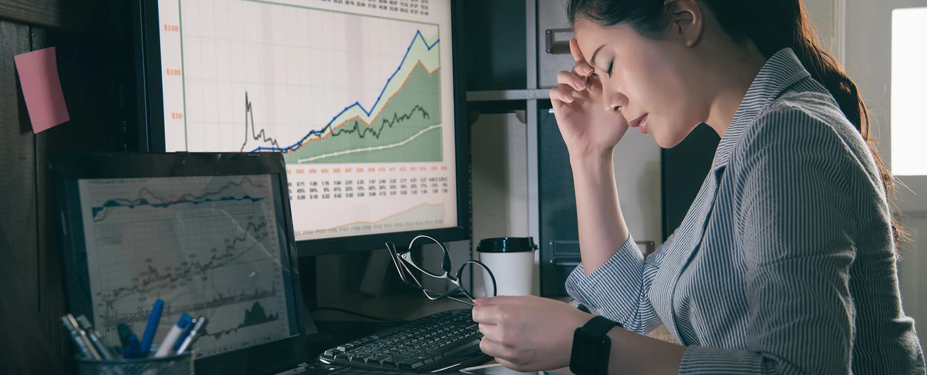Woman looking stressed while reviewing financial charts on computer screens at her desk.