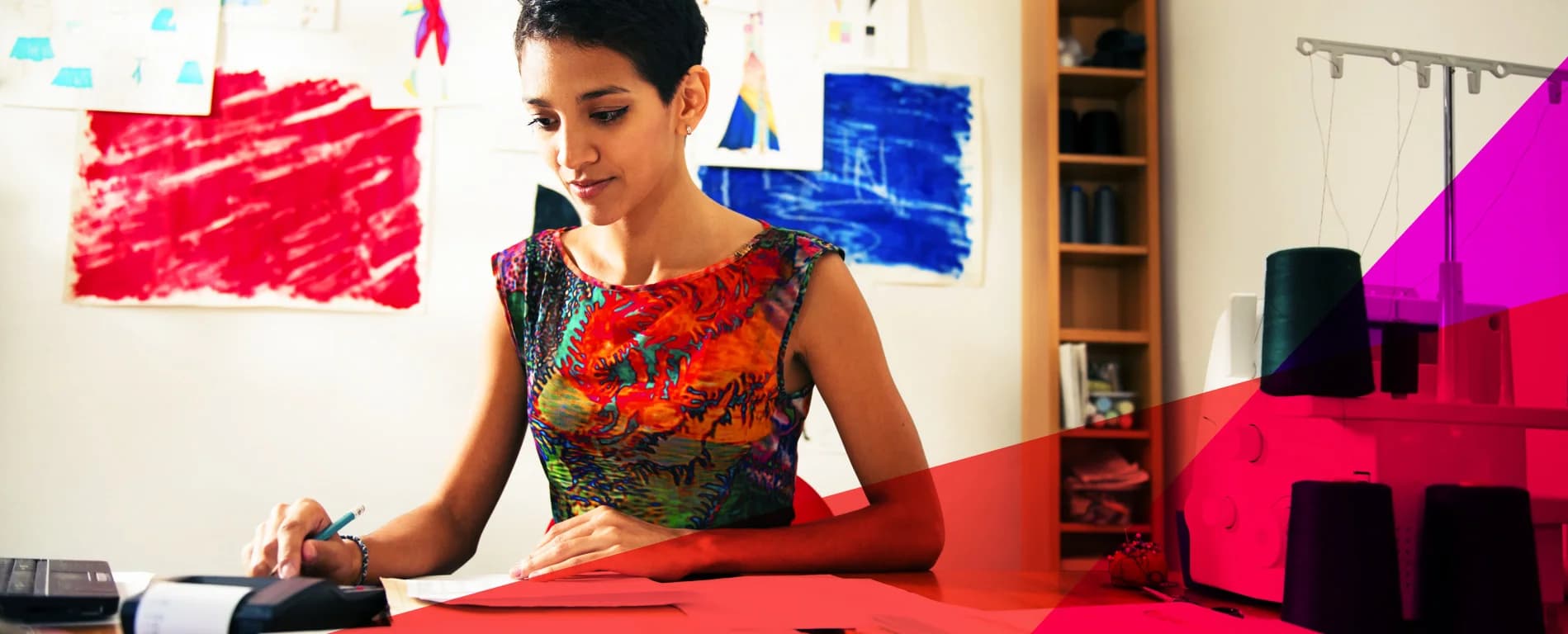 Woman working at a desk calculating expenses with a calculator and writing notes in a creative workspace with colorful artwork in the background.