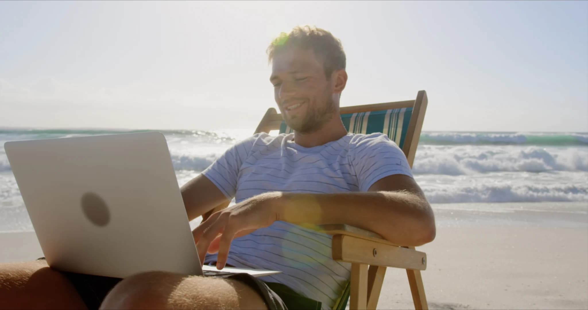 Man relaxing on a beach chair using a laptop by the ocean on a sunny day.
