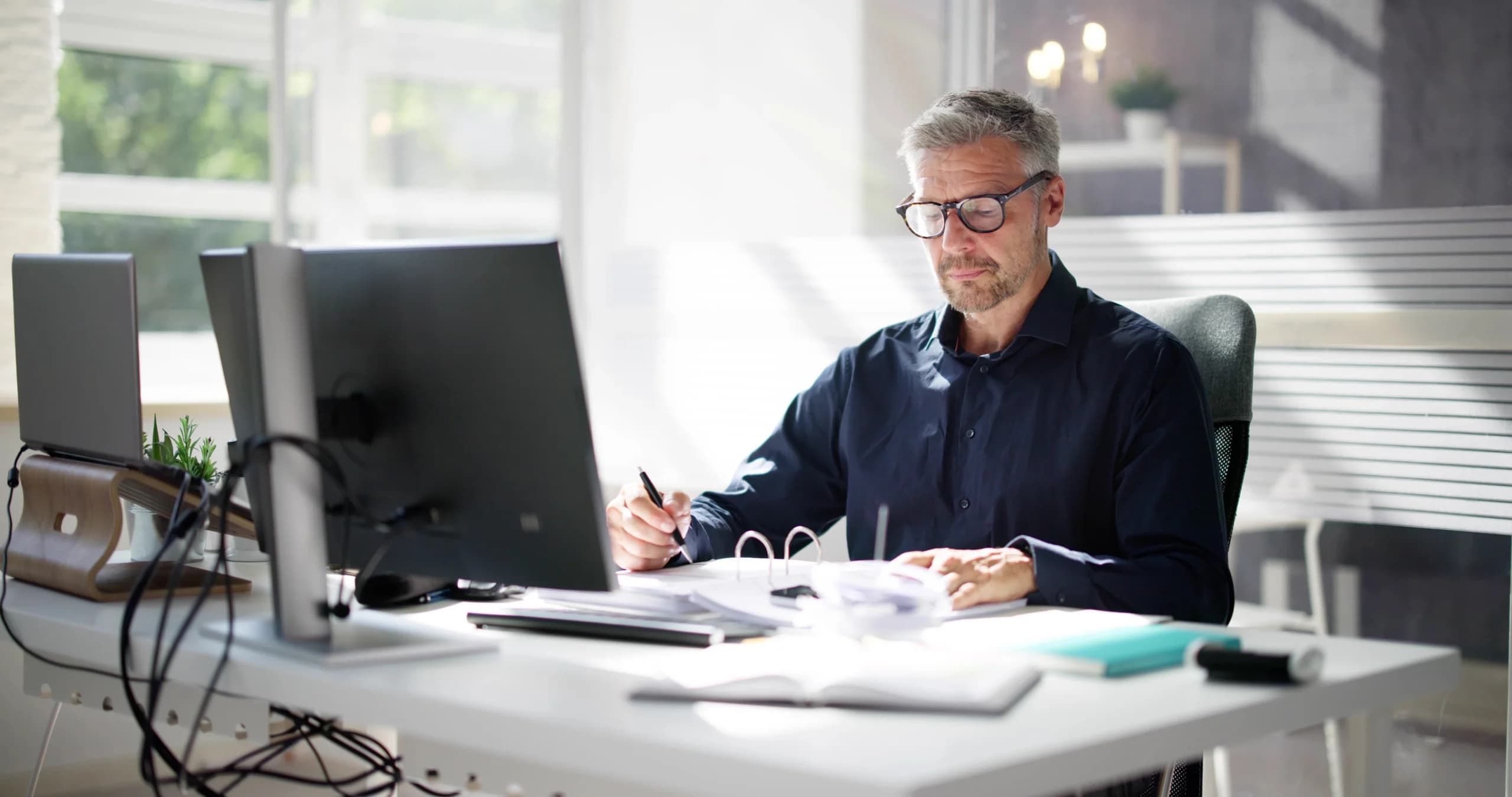 Man working at a desk reviewing documents and writing notes while using a computer in a modern office.