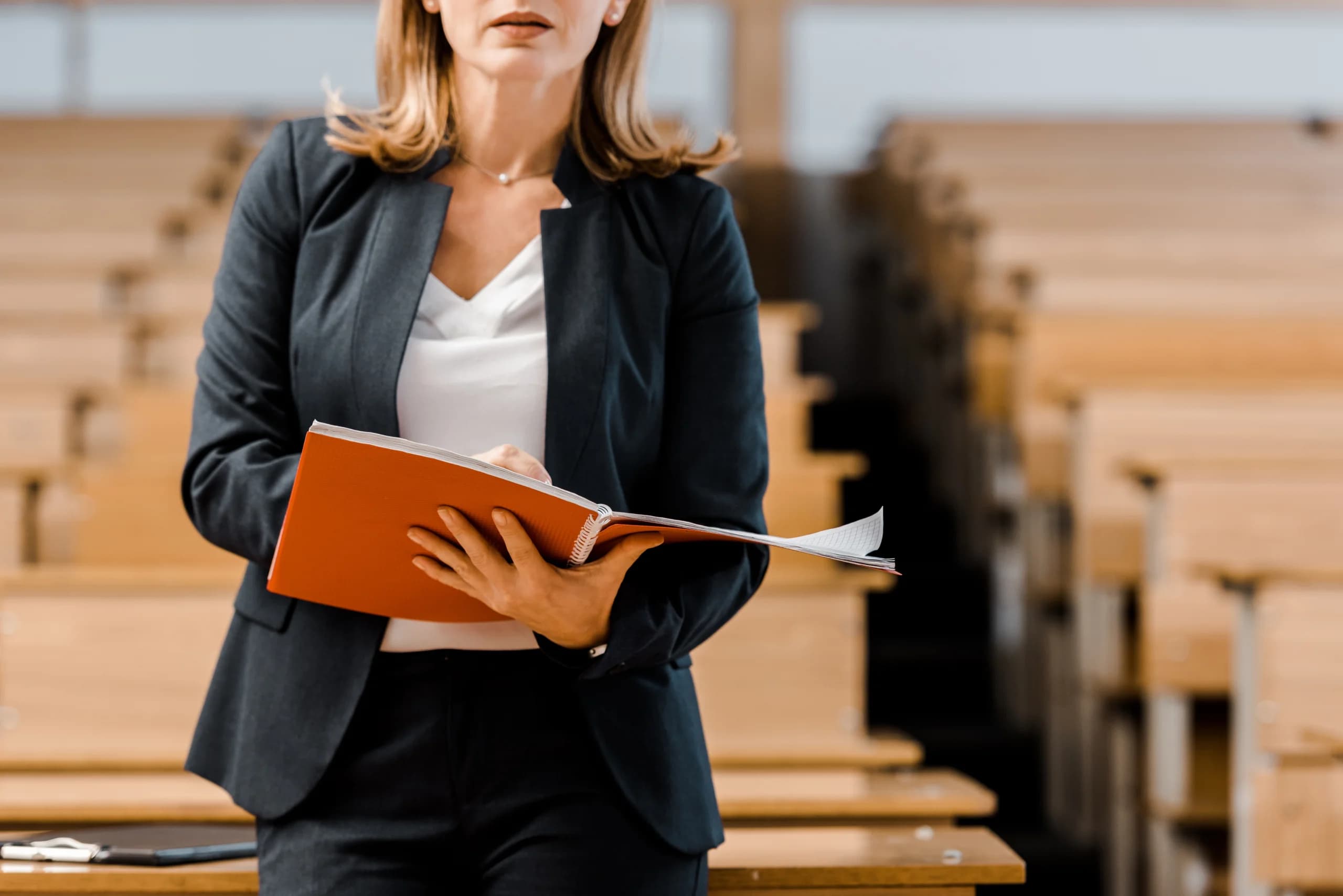 Woman holding a notebook standing in a lecture hall with empty seats, appearing prepared to teach or present.