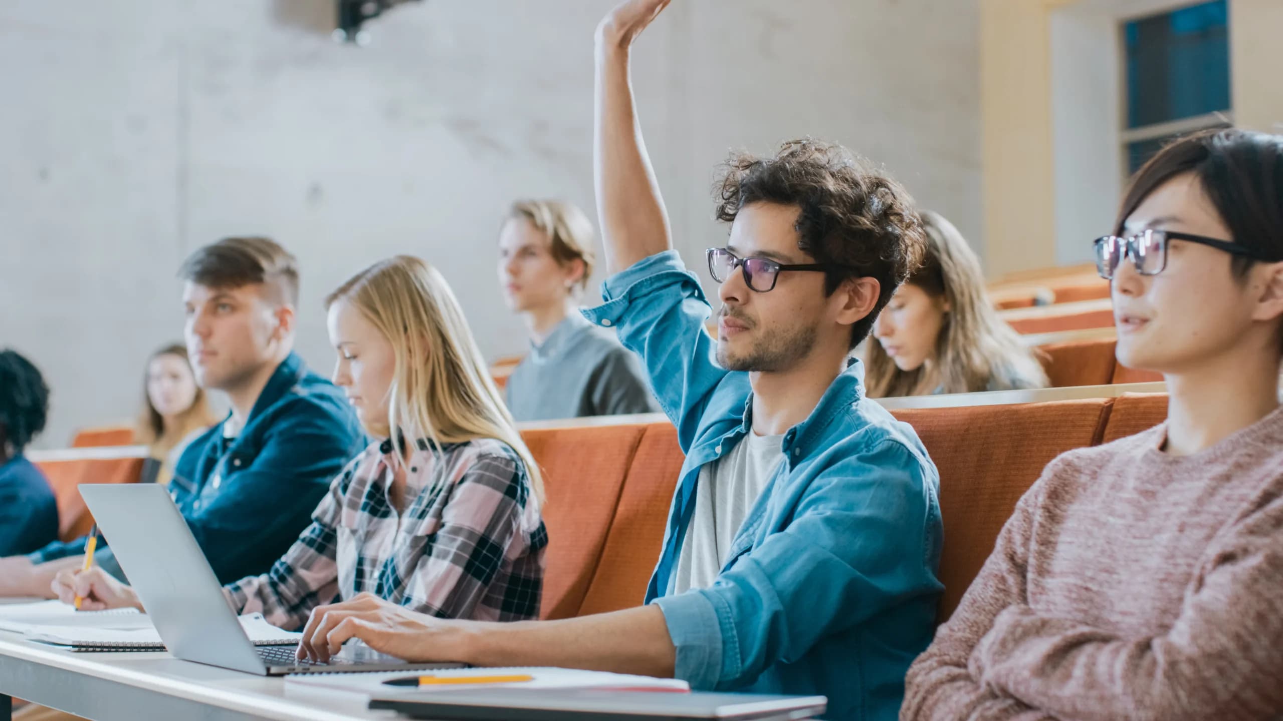 Students sitting in a lecture hall, with one male student raising his hand while others take notes and use laptops.