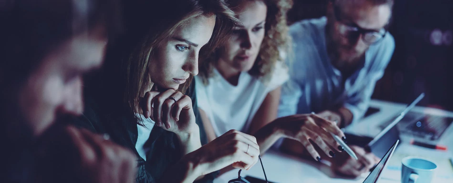 Three colleagues working closely together at a desk, focused on a laptop screen, discussing and analyzing information in a dimly lit office environment.