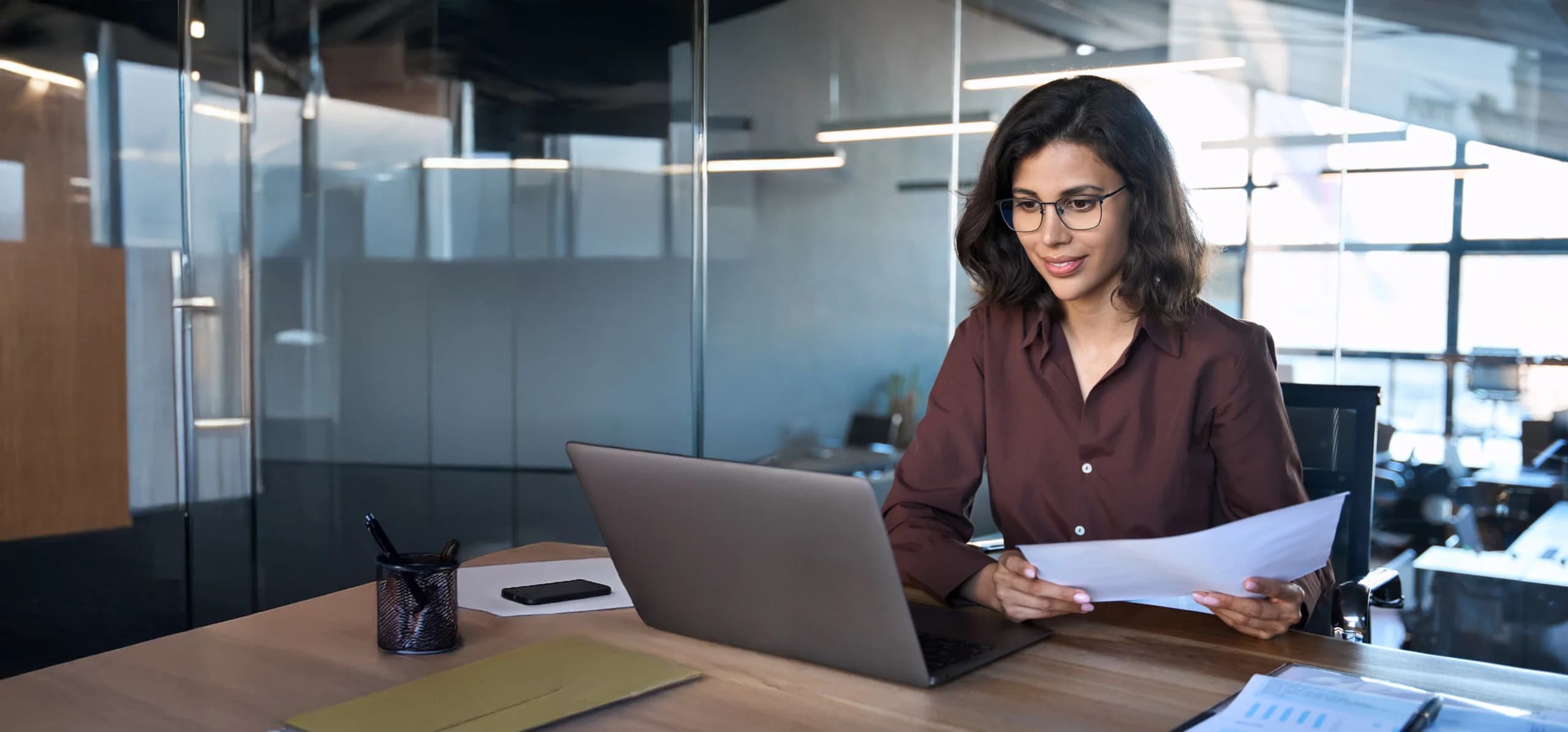 Businesswoman sitting at a desk in a modern office, reviewing documents while working on a laptop.
