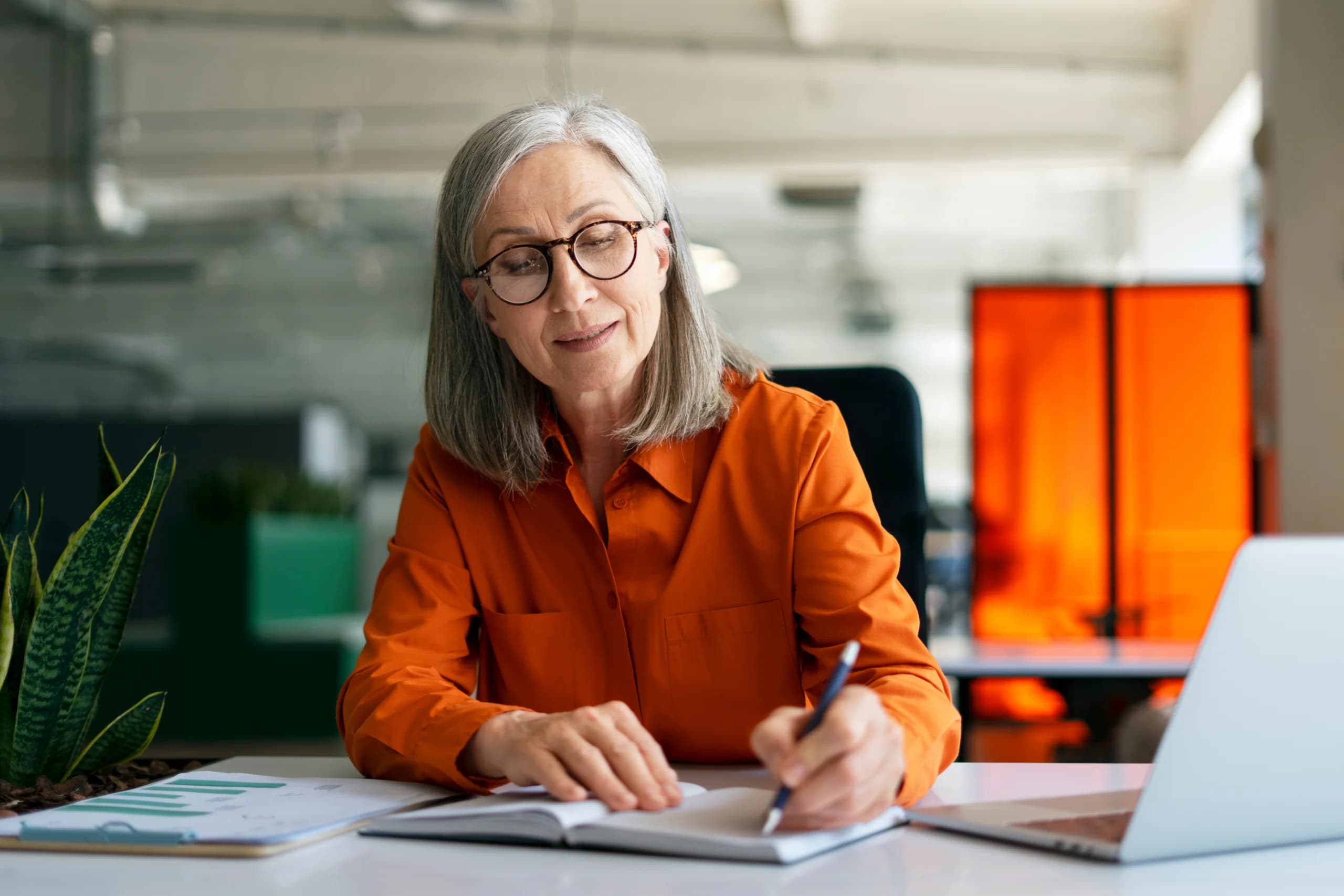 Older professional woman in an office, writing in a notebook at her desk with a laptop and documents nearby.