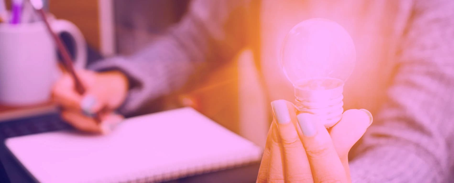 Hand holding a glowing light bulb in the foreground while another person writes in a notebook beside a laptop in a warm, blurred setting.