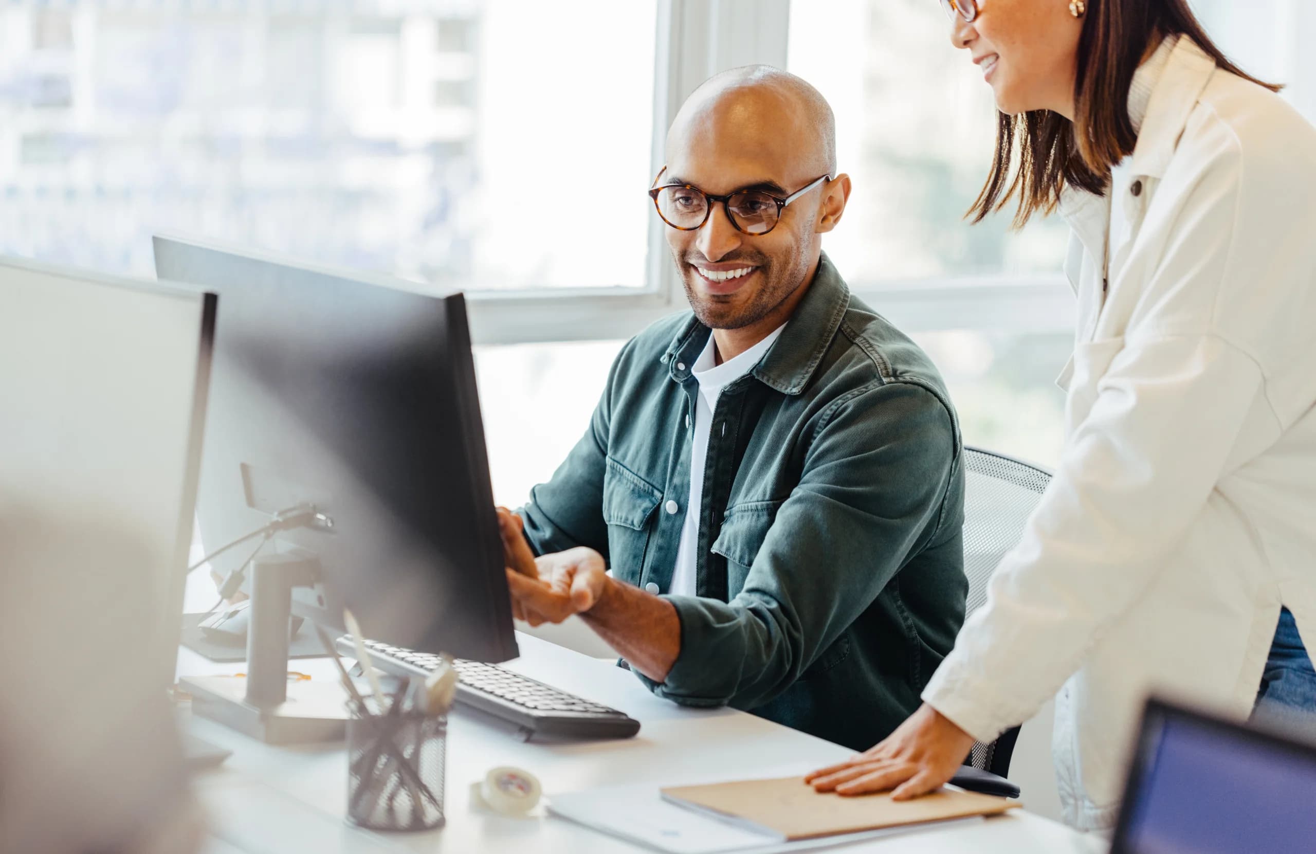 Two colleagues smiling and collaborating at a desk, looking at a computer screen in a bright office environment.