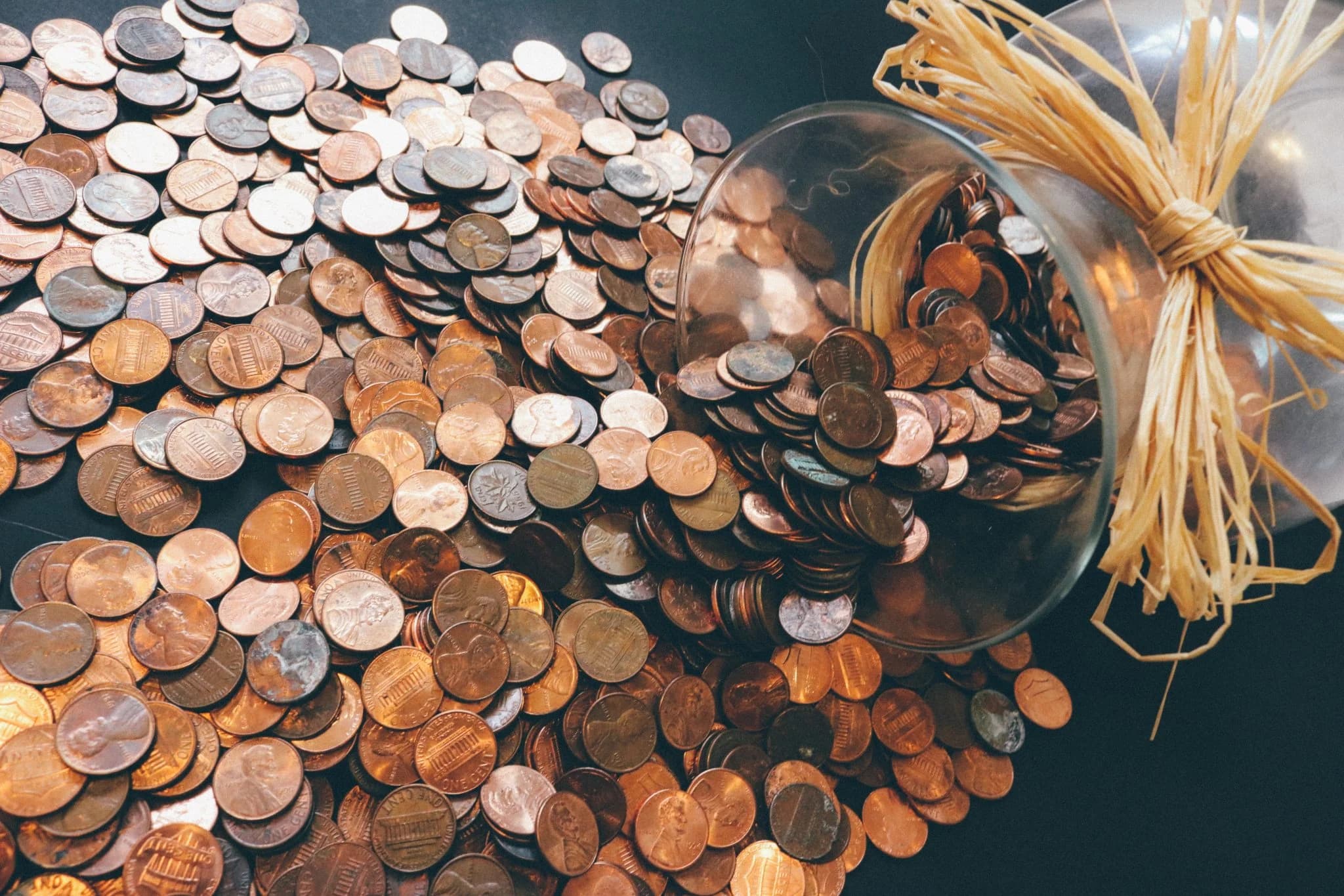 Spilled glass jar filled with coins lying on its side, with numerous coins scattered across a dark surface.