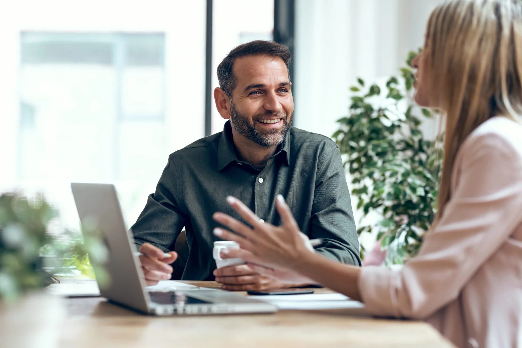Two colleagues having a friendly conversation at a desk with a laptop, one holding a coffee cup in a bright office setting.