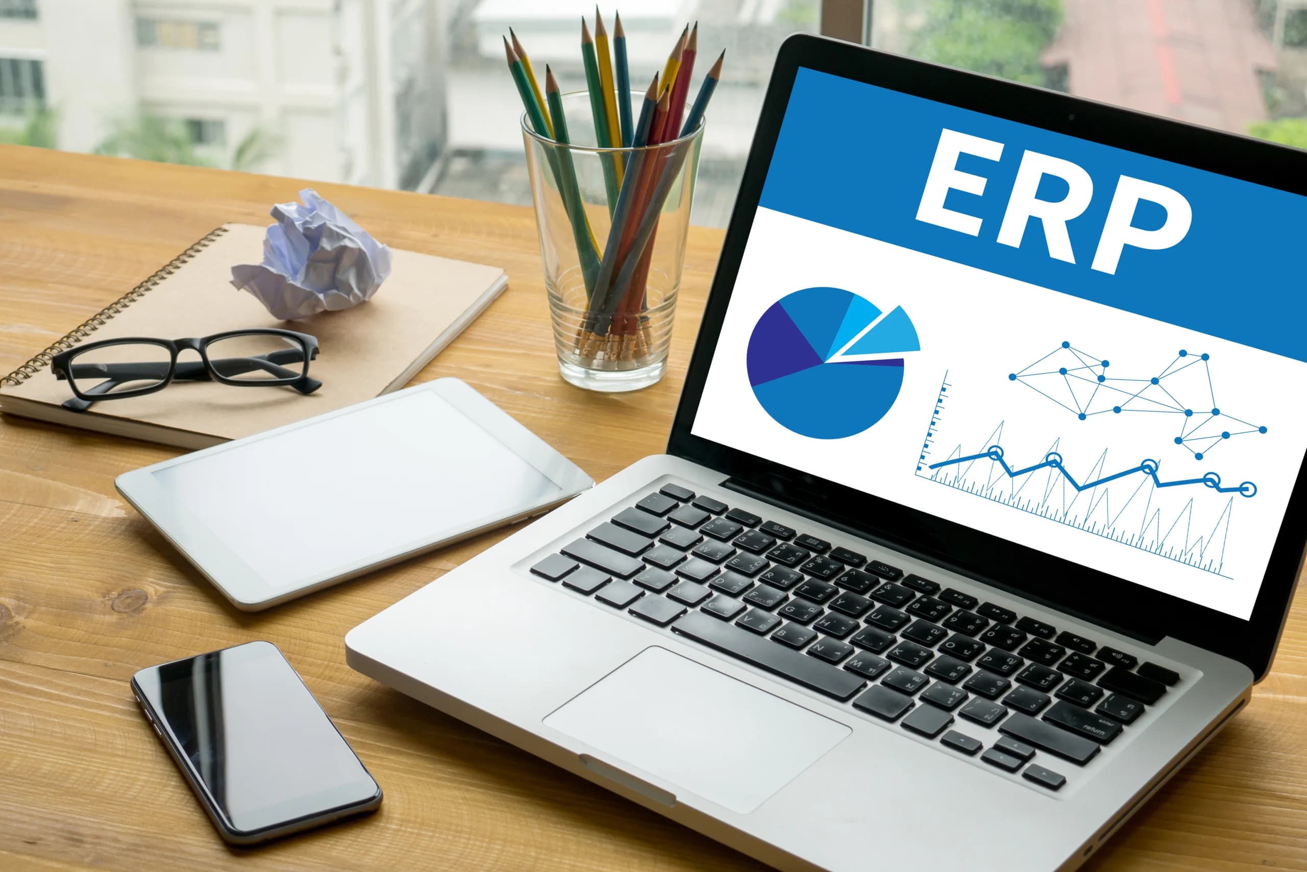 Laptop displaying an ERP dashboard with charts and graphs on a desk alongside a tablet, smartphone, notebook, glasses, and a cup of pencils.