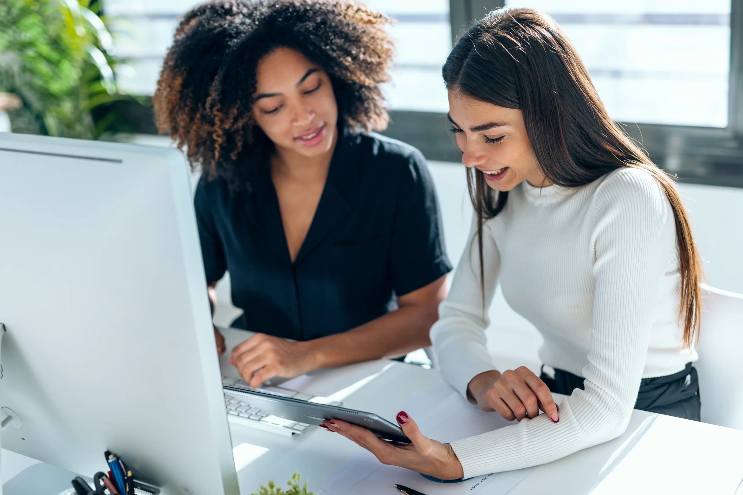 Two colleagues collaborating at a desk, looking at a tablet together in a bright office environment.