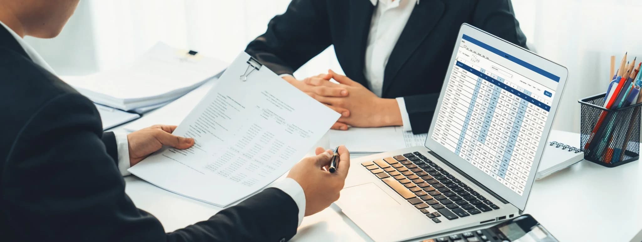 Business professionals reviewing financial documents at a desk with a laptop displaying a spreadsheet.