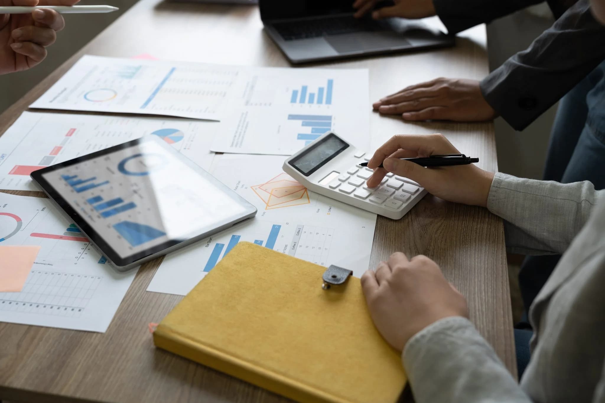 Person using a calculator on a desk with financial charts, a tablet, and documents while others collaborate in the background.