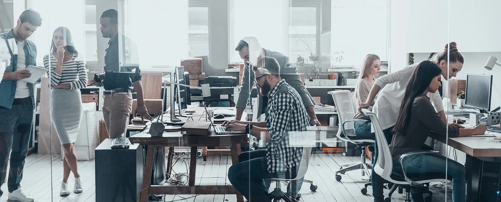 Open-plan office with multiple colleagues collaborating at desks, using computers and discussing work in a modern workspace.