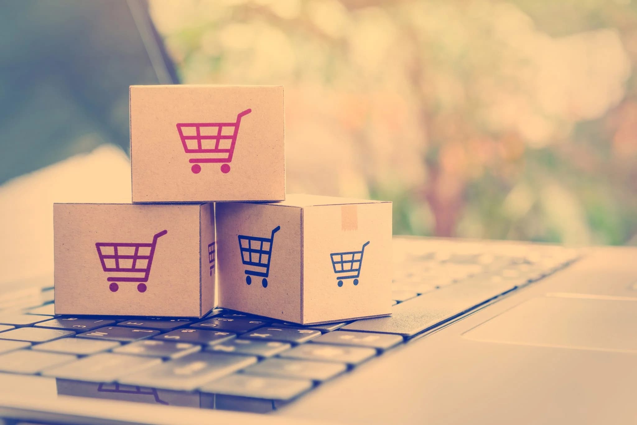 Stacked cardboard boxes with shopping cart icons placed on a laptop keyboard, representing e-commerce and online shopping.