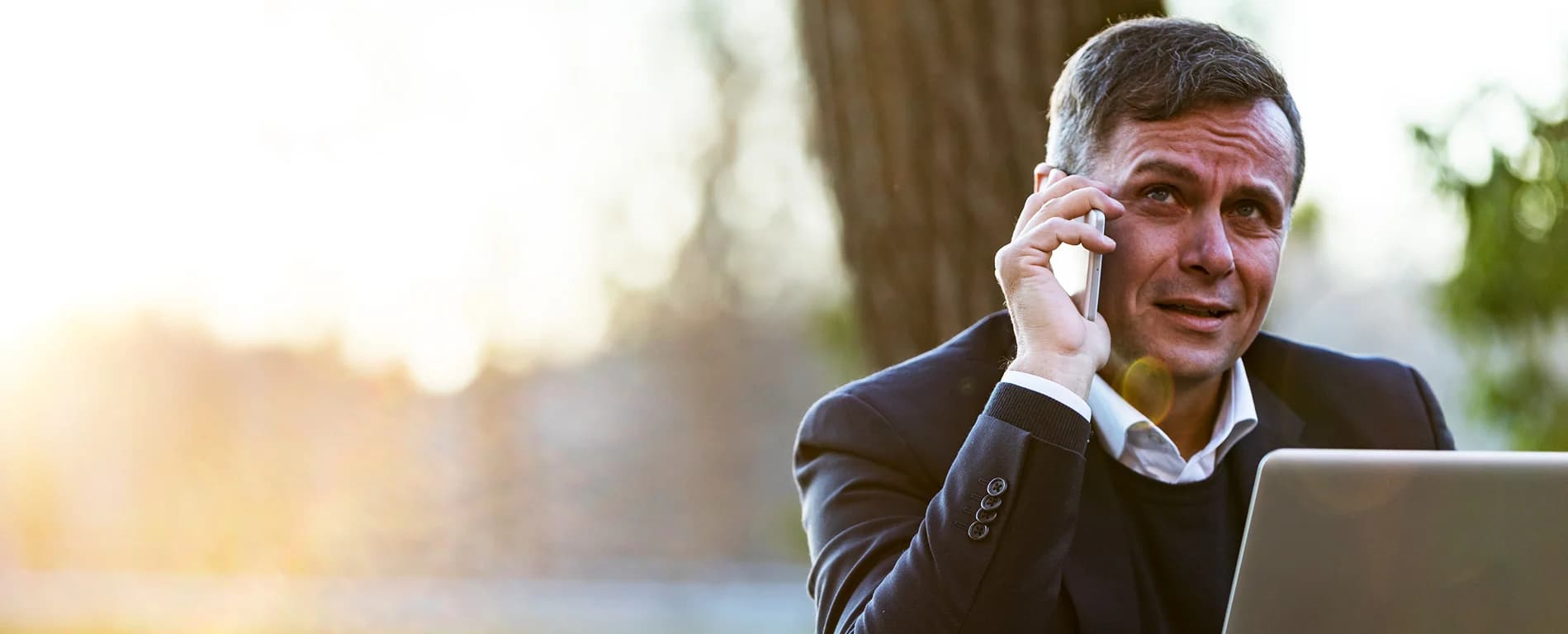 Businessman talking on a smartphone while working on a laptop outdoors in a sunlit setting.