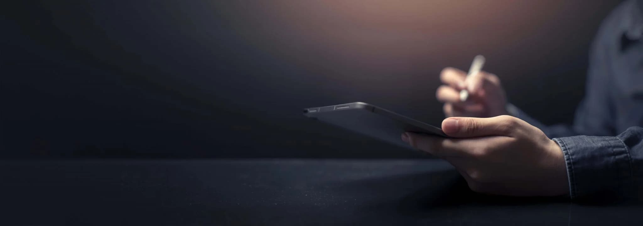 Close-up of a person using a tablet with a stylus against a dark background, suggesting digital work or design