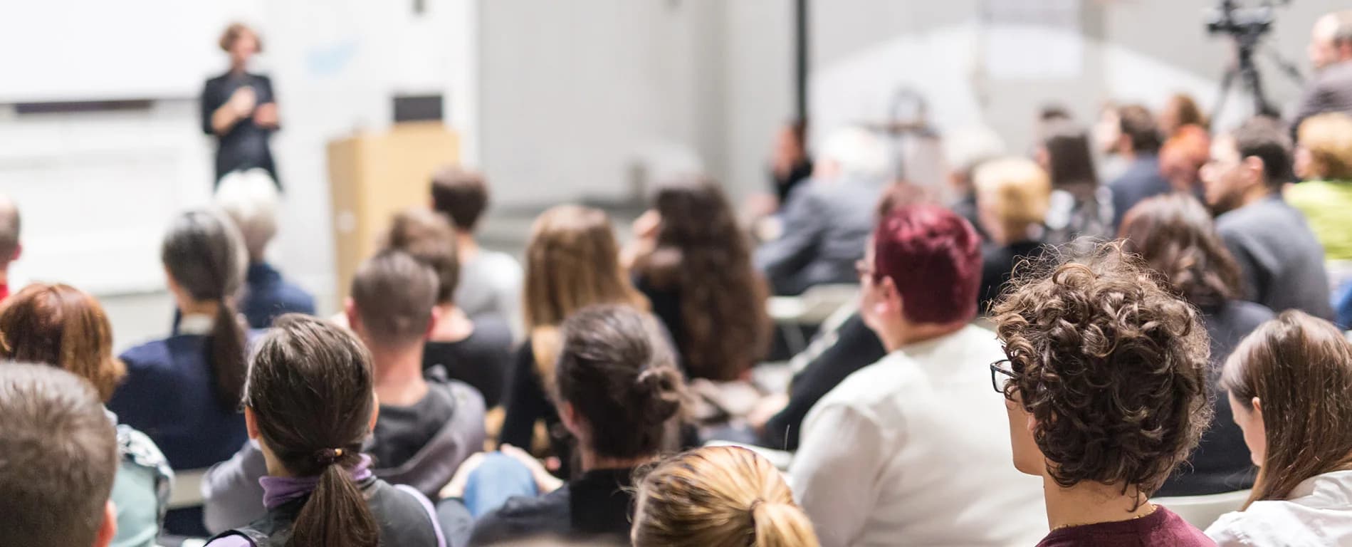 Audience attending a presentation or lecture, facing a speaker at the front of a conference room