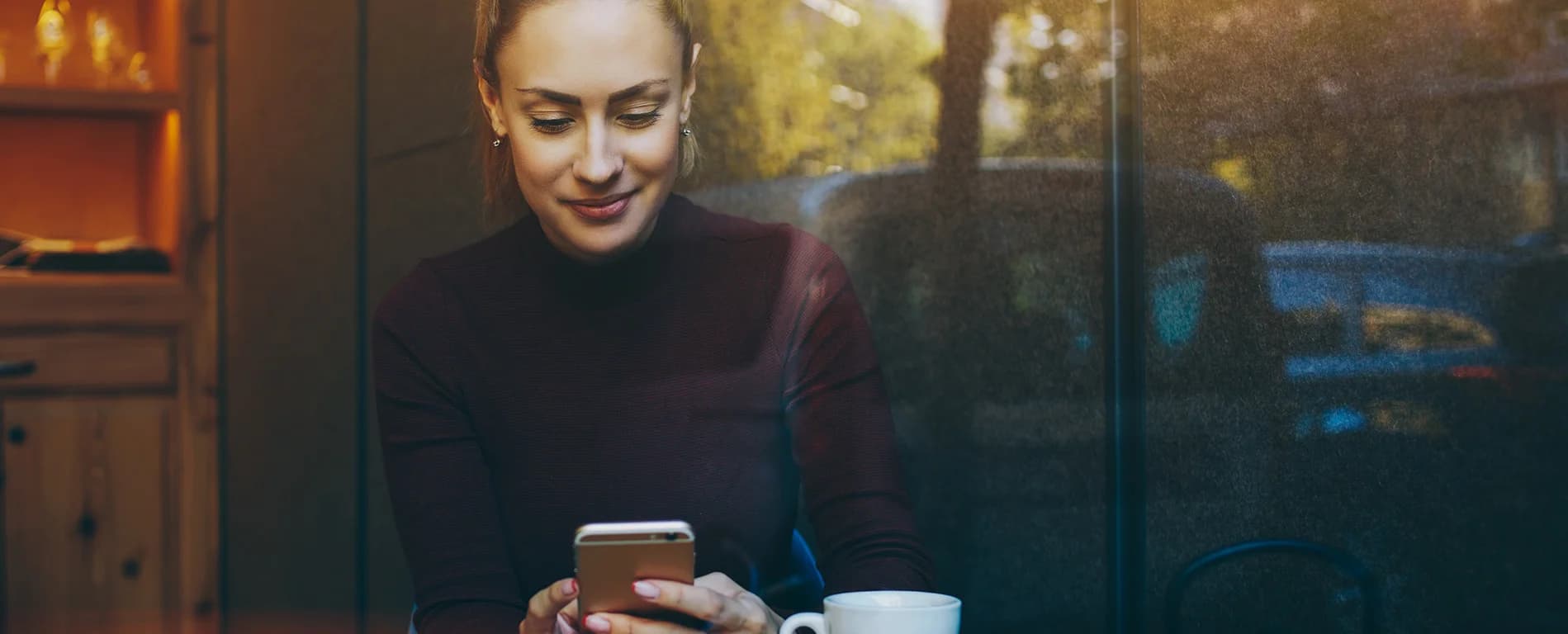 Woman sitting by a window in a café, smiling while using her smartphone with a cup of coffee nearby