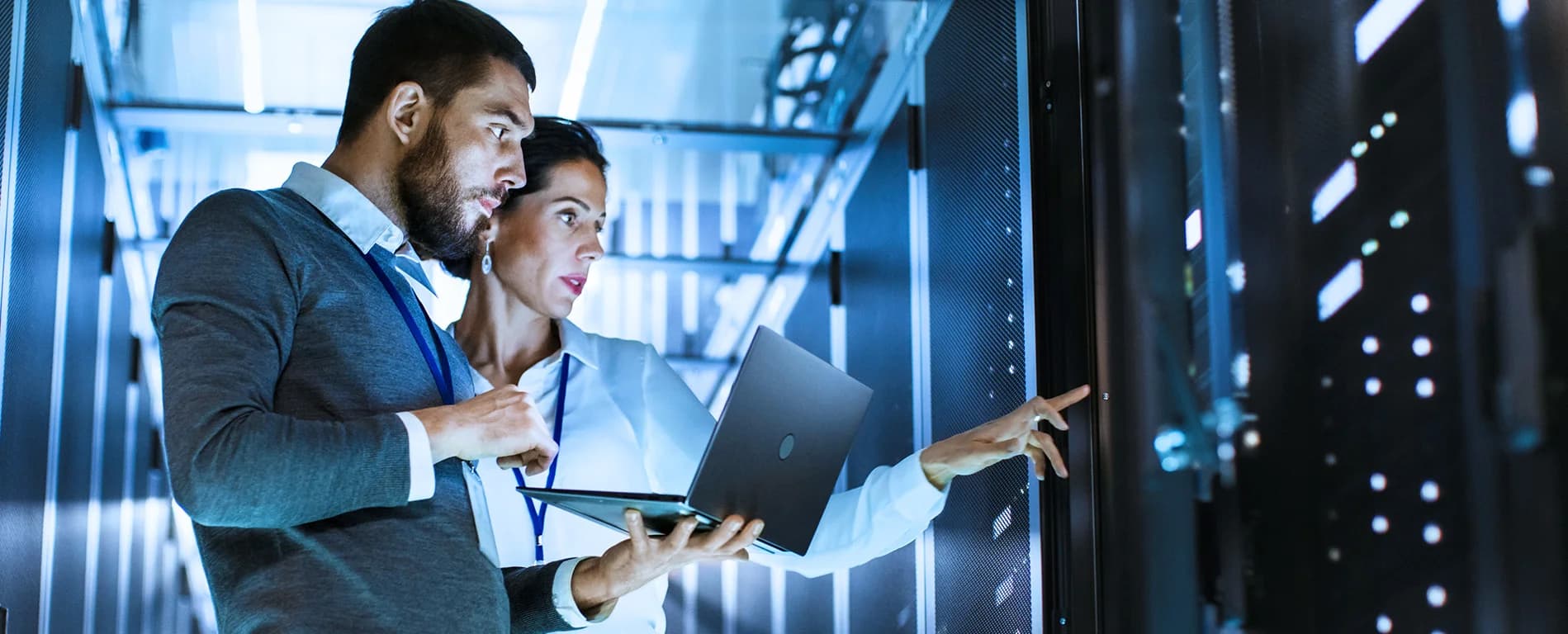 Two IT professionals working together in a server room, looking at a laptop and checking equipment.