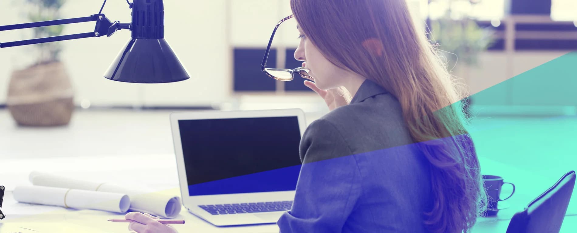 Woman working at a desk with a laptop, holding her glasses and looking focused.