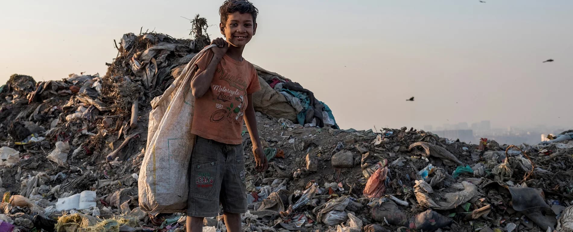 Boy carrying a sack over his shoulder, standing in a large landfill area.