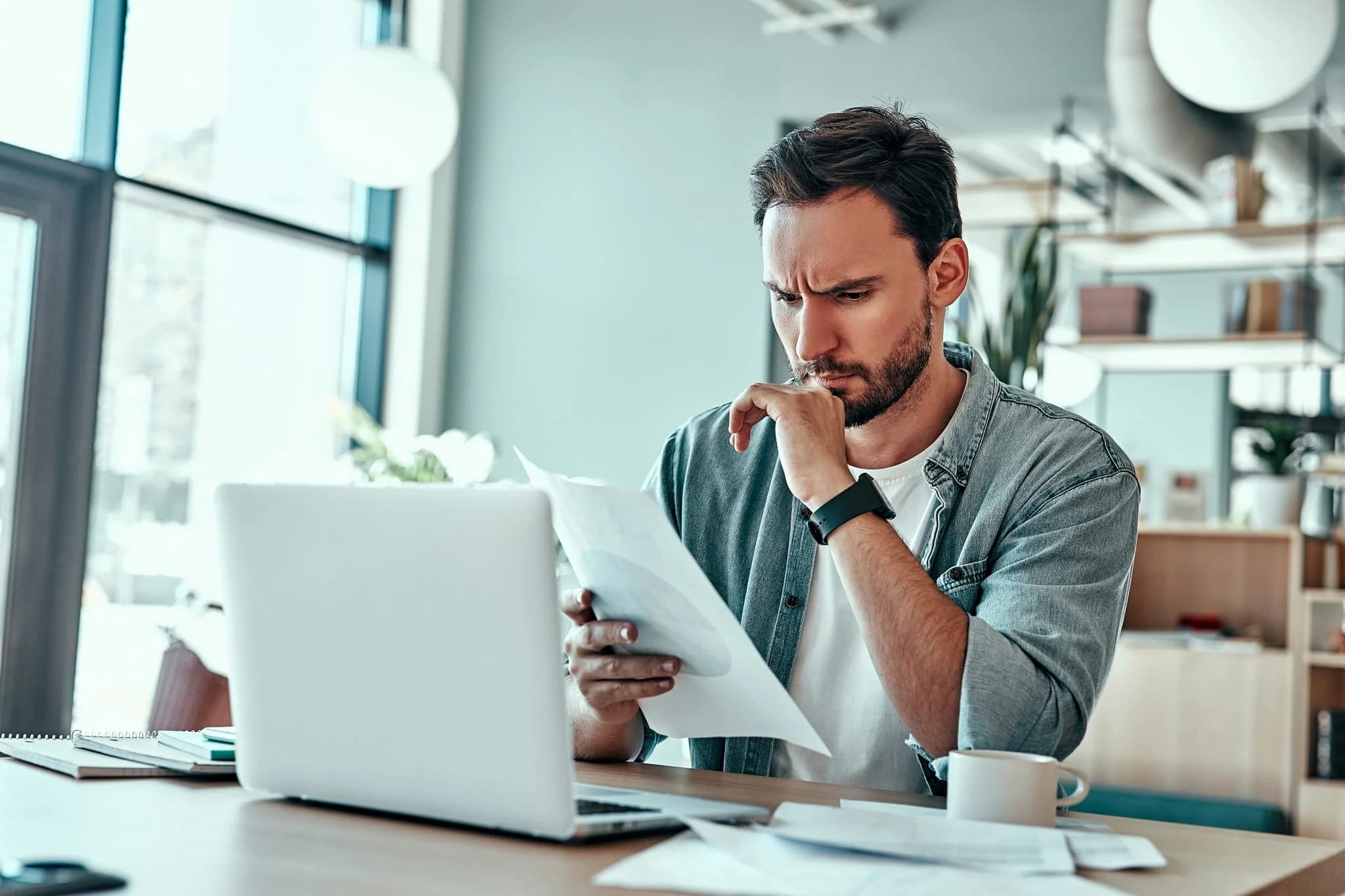 Man sitting at a desk reviewing documents with a concerned expression, using a laptop in a modern office setting.