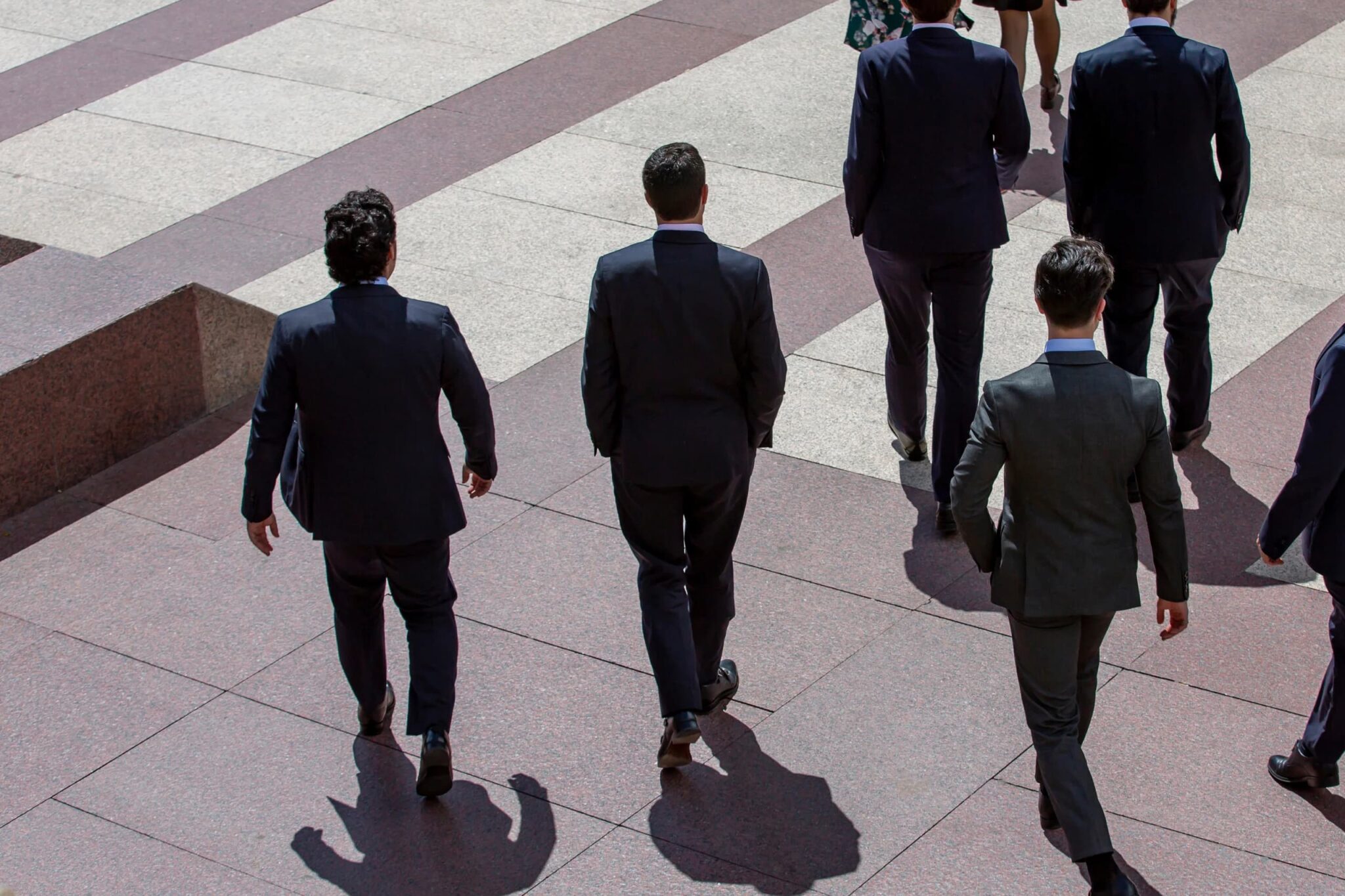 Group of business professionals in suits walking across an urban plaza, viewed from above.