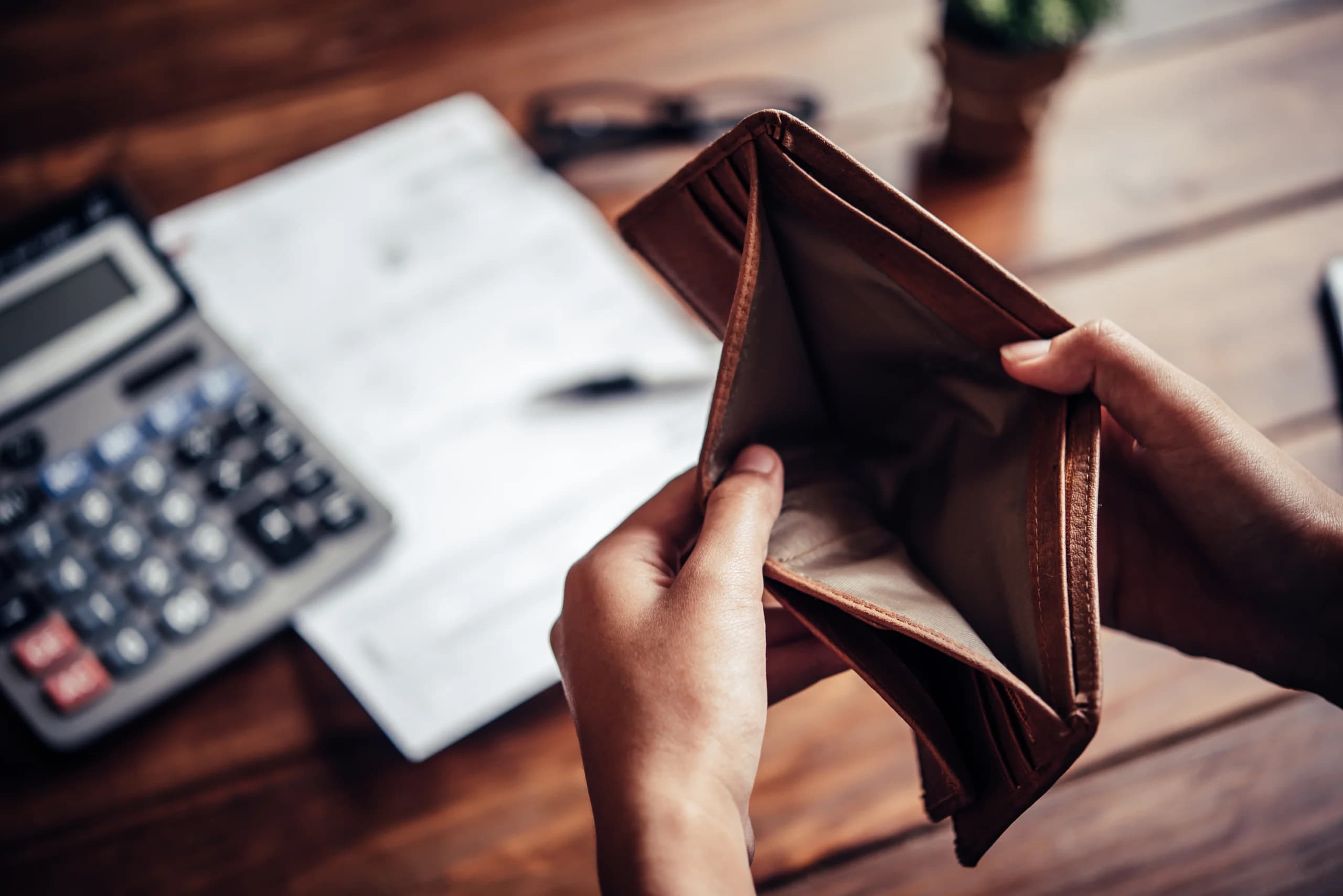 Person holding an empty wallet at a desk with a calculator and bills, suggesting financial stress or lack of funds.
